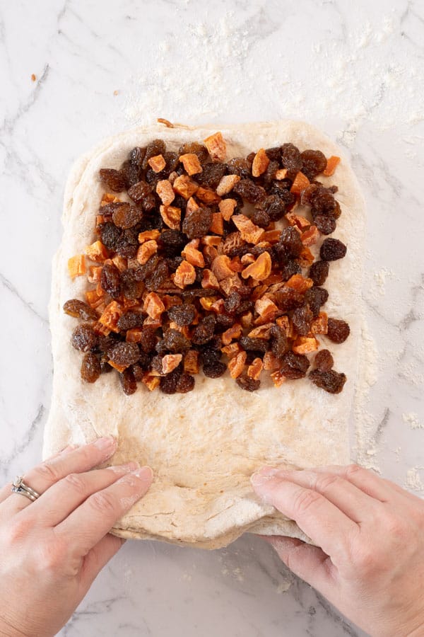 a rectangle of bread dough on a marble benchtop. The dough has dried fruit and nuts scattered over the top, which are being folded into it with two hands visible.