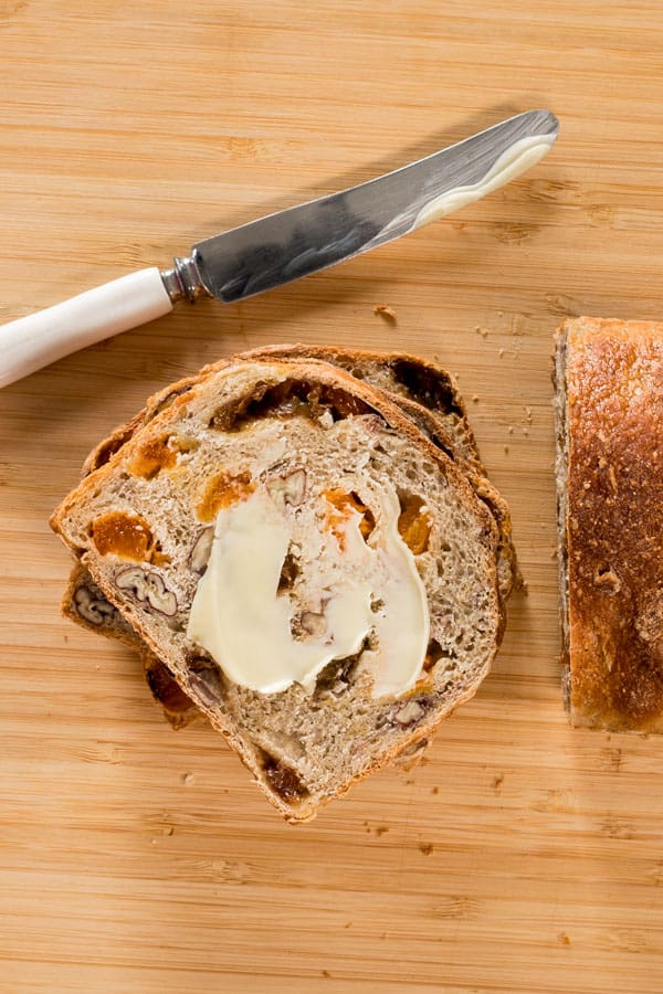 a wooden chopping board with a small stack of fruit and nut loaf slices viewed from above. The top slice has a smear of butter across it, with the butter knife next to the bread on the board.