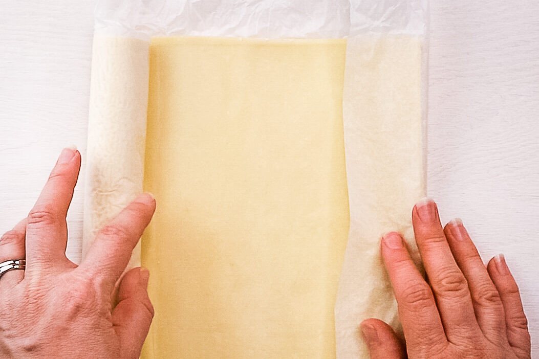 A woman's hands, with a silver ring on her left ring finger, roll out a piece of puff pastry onto parchment paper