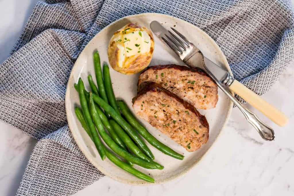 A white ceramic plate with two slices of steam oven meatloaf, a buttered baked potato and cooked green beans. A knife and fork lay alongside the plate.