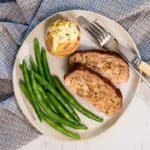 A white ceramic plate with two slices of steam oven meatloaf, a buttered baked potato and cooked green beans. A knife and fork lay alongside the plate.