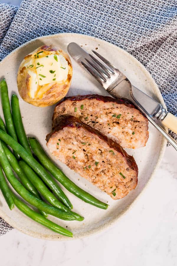 Steam oven meatloaf with baked potato and green beans, on a white plate with a black and white checked napkin underneath it.