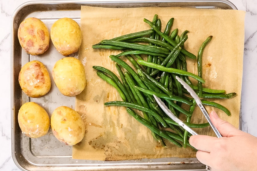 A metal sheet pan with six baked potatoes and a pile of green beans. The beans are being picked up with a pair of tongs.