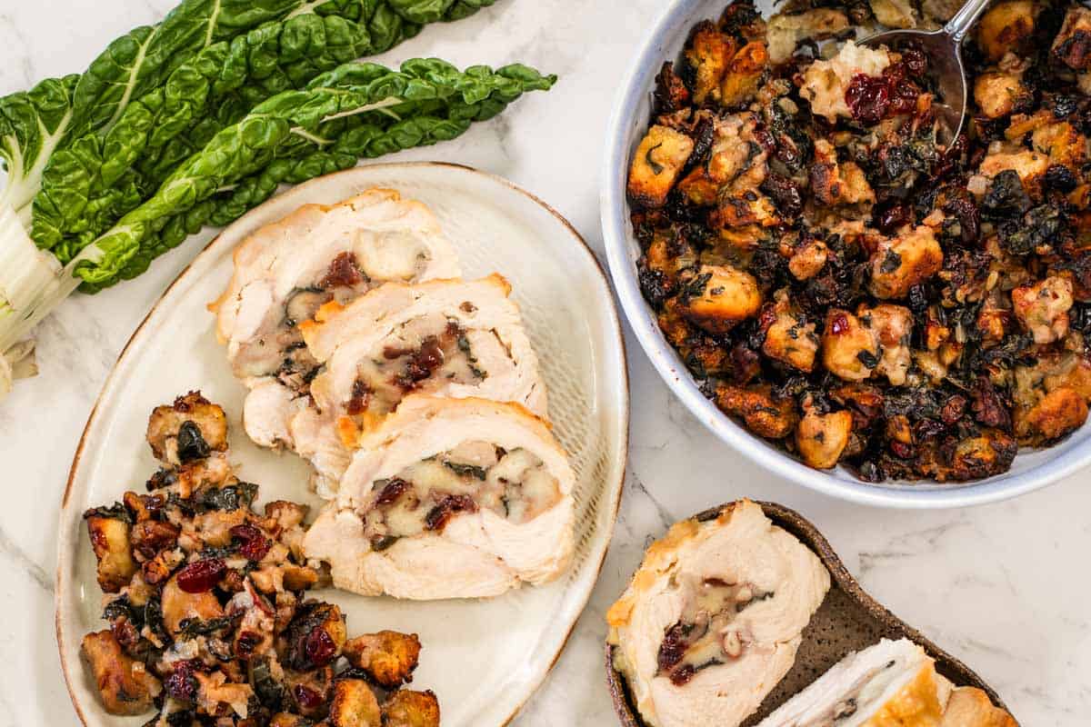 A table spread with swiss chard leaves, rolled and stuffed turkey breast, and cranberry pecan stuffing. The crockery is white and the table is marble.