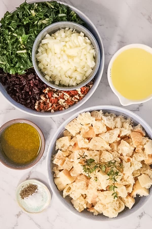 Ingredients for cranberry pecan turkey stuffing, set out in bowls. Swiss chard; onion, chicken stock, cubed bread, thyme, salt, pepper, oil, pecans and cranberries. 