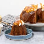 A sticky gingerbread steamed pudding on a large footed plate. The pudding has a piece cut out which can be seen on a plate in front; butterscotch sauce and whipped cream garnish the pudding.