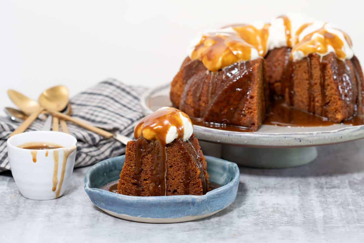 A sticky gingerbread steamed pudding on a large footed plate. The pudding has a piece cut out which can be seen on a plate in front; butterscotch sauce and whipped cream garnish the pudding.