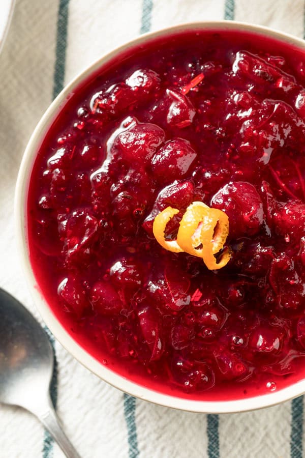 a green and white striped tablecloth with a bowl of sous vide cranberry sauce viewed from the top down. A curl of orange zest is on top of the sauce.