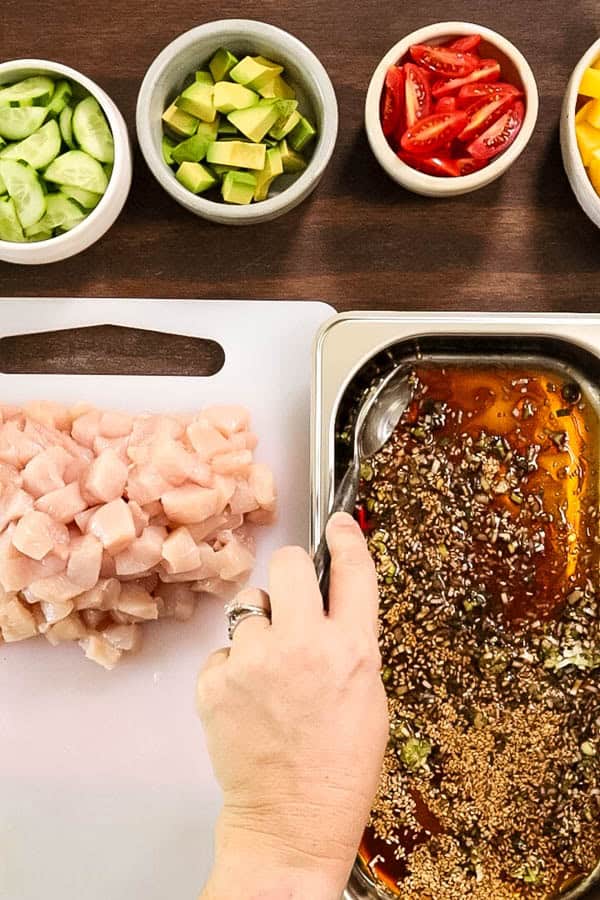 a hand stirs soy sauce marinade for chicken, to make chicken poke bowls. Alongside is chopped chicken breast and bowls of brightly colored vegetables and fruits for the bowls. 