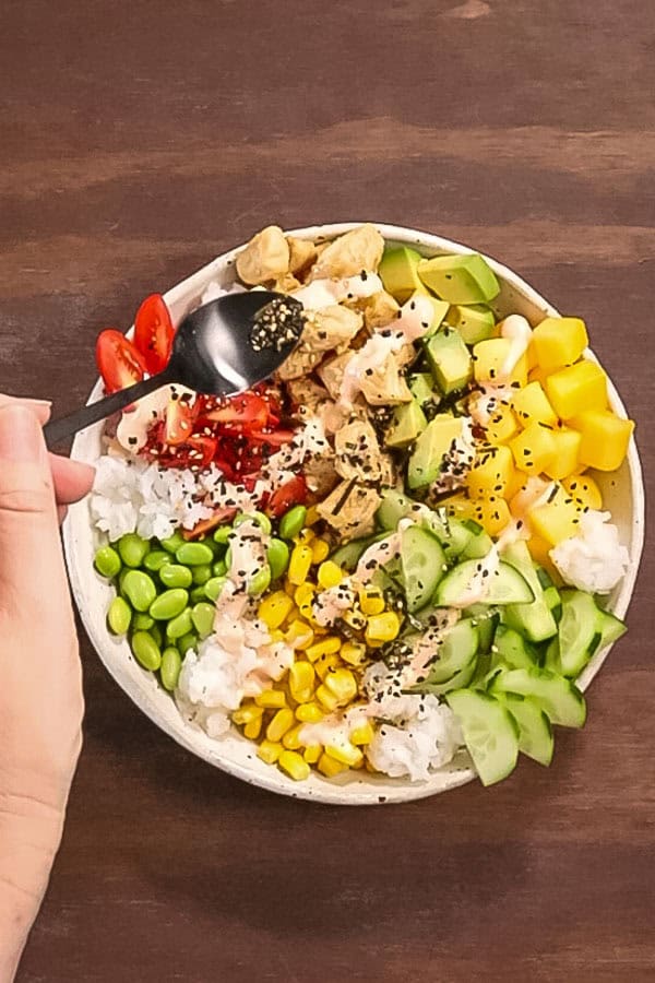 a hand holds a black metal teaspoon, sprinkling furikake seasoning over the top of a chicken poke bowl with vegetables and spicy mayonnaise.