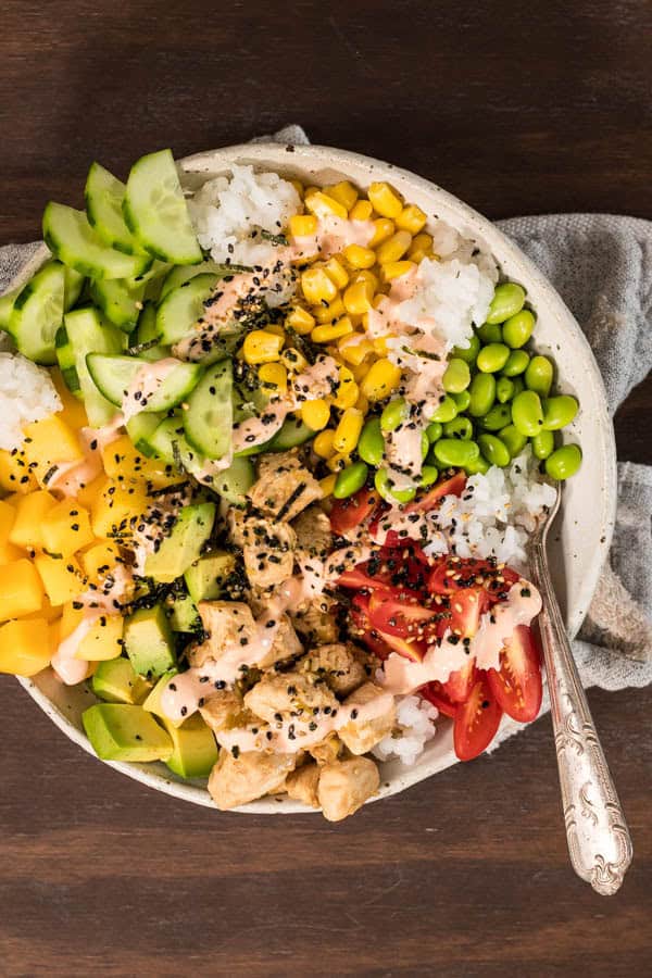 A finished steamed chicken poke bowl, with colorful fruits and vegetables, spicy mayo and furikake seasoning on top of sushi rice. 