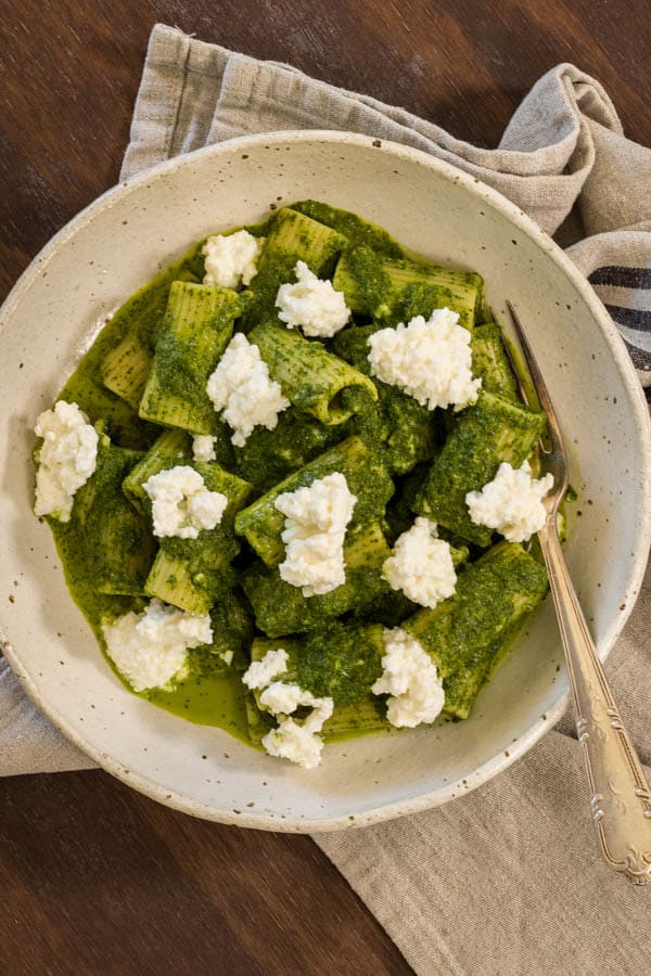 a bowl viewed from above, filled with green kale sauce coated pasta and dots of ricotta cheese