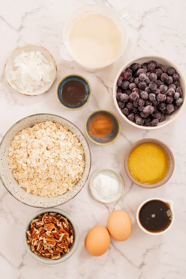 Ingredients for blueberry pecan baked oatmeal laid out on a marble countertop. Each ingredient is in a small bowl or dish. 