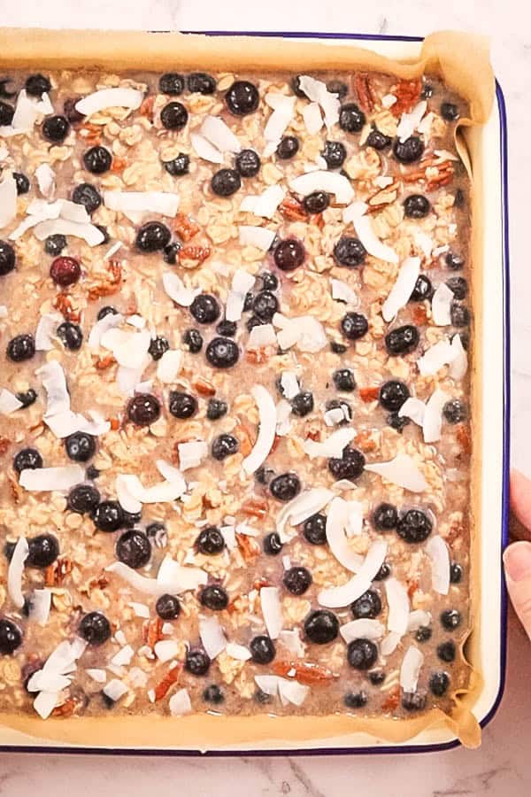 A pan of baked oatmeal batter, about to go into the oven. There are blueberries and flakes of coconut visible on top of the oats and nuts. 