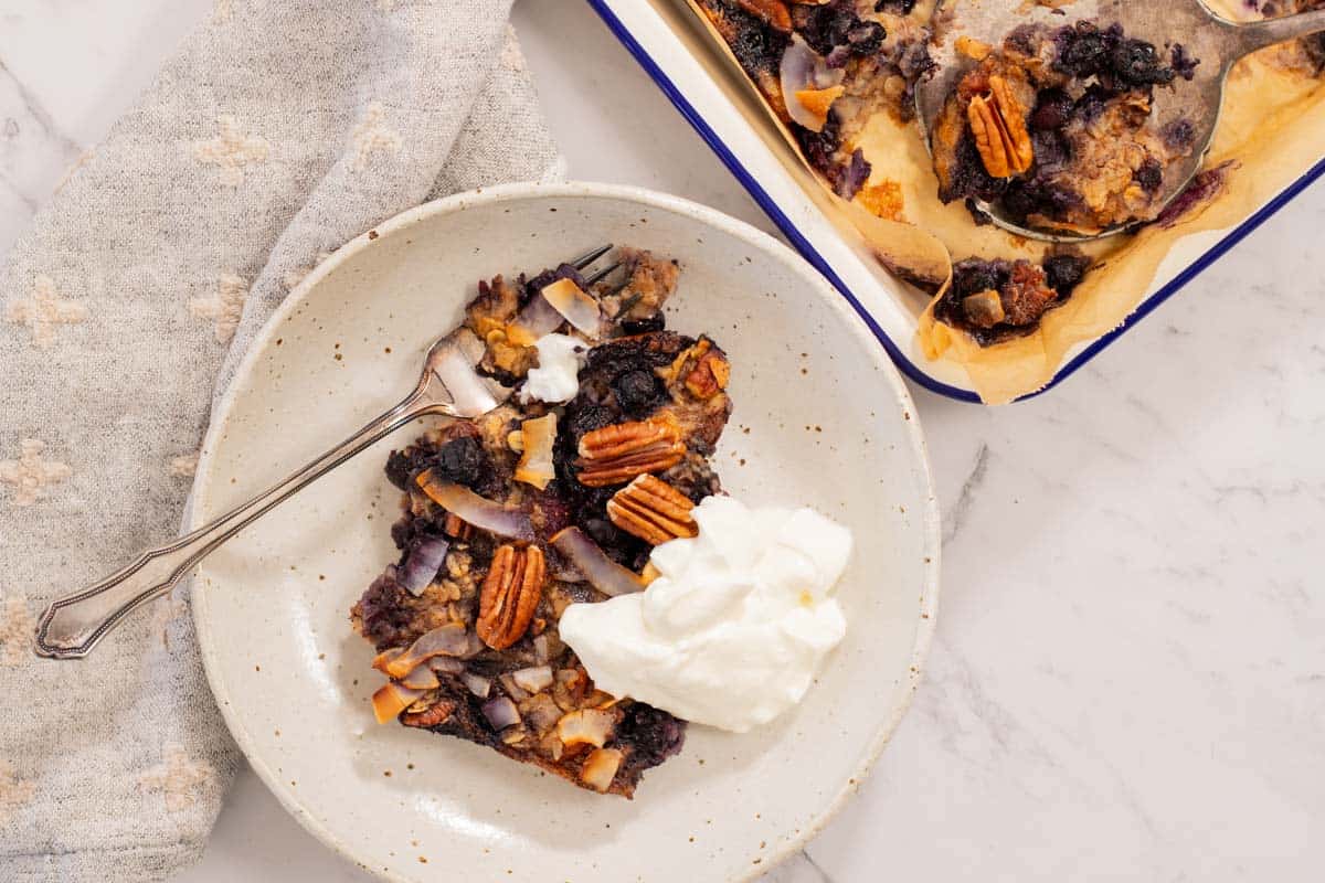 Blueberry and pecan baked oatmeal, scooped onto a white plate and served with a dollop of thick yogurt. A fork sits on the plate, and a beige napkin alongside.