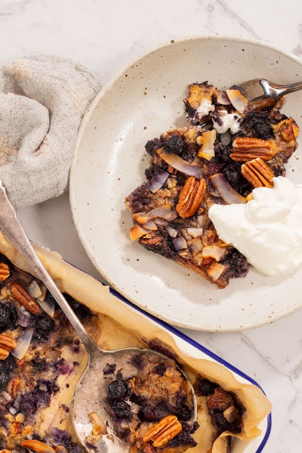 A top down view of baked blueberry and pecan oatmeal, served in a white ceramic dish with thick yogurt alongside. There is a beige napkin next to the dish. 