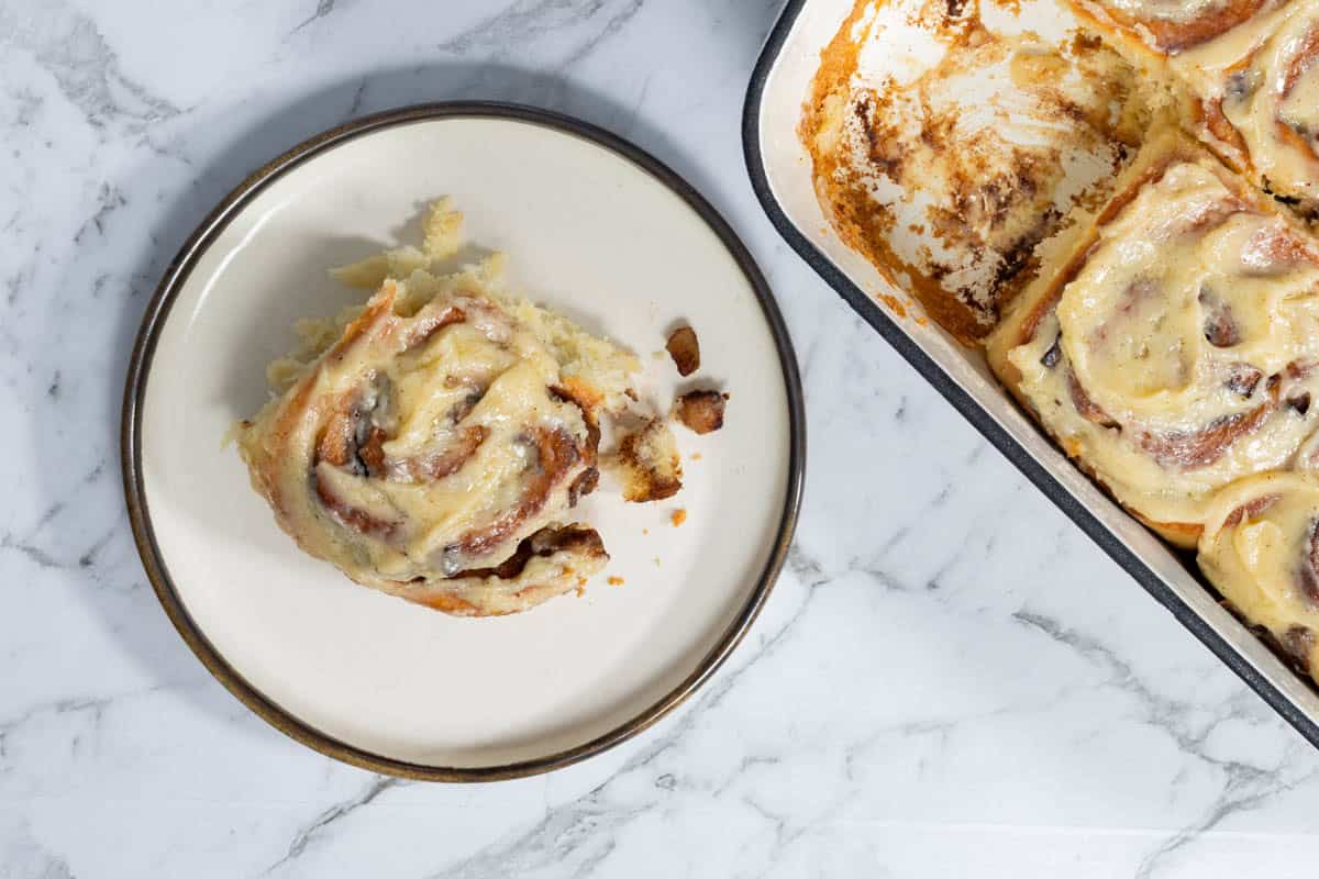 Top-down view of a soft, glazed apple cinnamon roll served on a round plate next to a baking dish filled with frosted rolls.