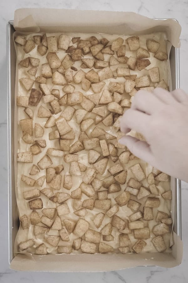 Hand scattering cinnamon-coated apple chunks over cake batter in a lined baking pan.