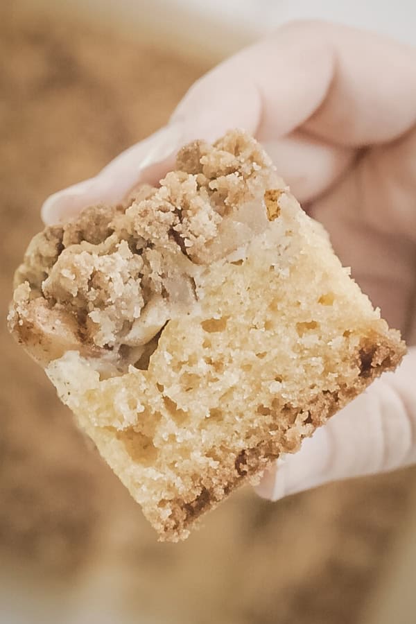 A close-up of a hand holding a slice of apple crumb cake, showing its soft, fluffy interior and buttery crumb topping.