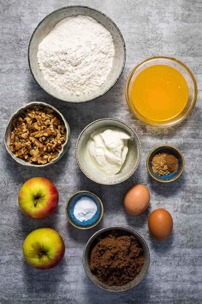 Ingredients laid out for apple walnut bread in small bowls. Flour, butter, walnuts, yoghurt, spices, apples, eggs, baking soda, brown sugar.