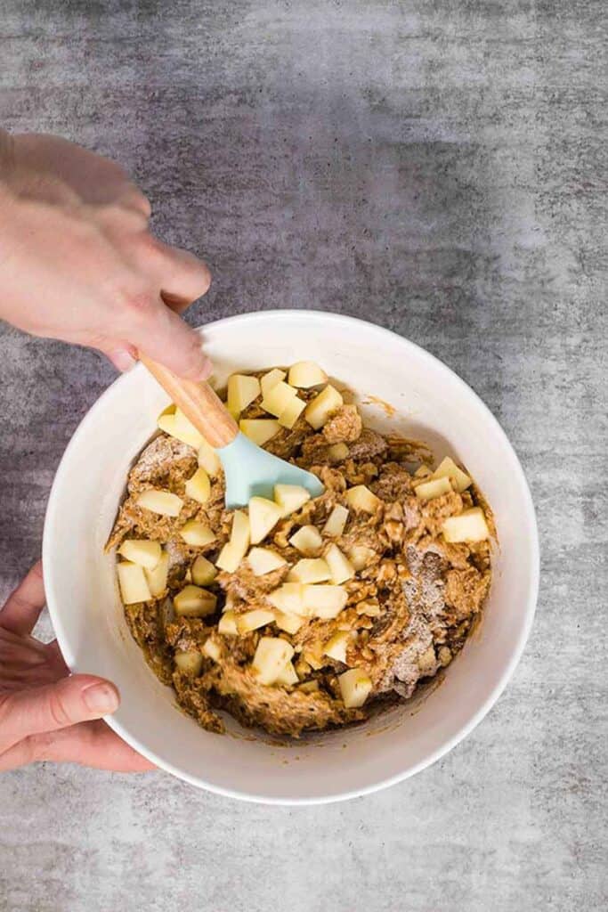 A white bowl and blue spatula, stirring chopped apples and walnuts into apple walnut loaf batter.