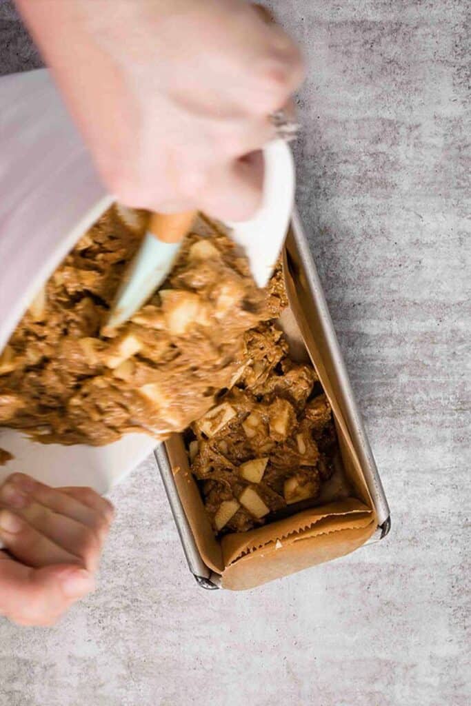 Scraping apple walnut bread batter from a white mixing bowl into a lined loaf pan.