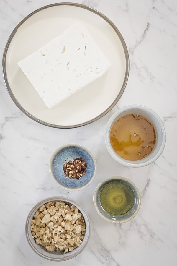 Ingredients for baked feta including a block of feta cheese, honey, walnuts, olive oil, and red pepper flakes.