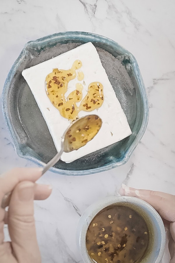 Drizzling honey and red pepper mixture over a block of feta in a baking dish.