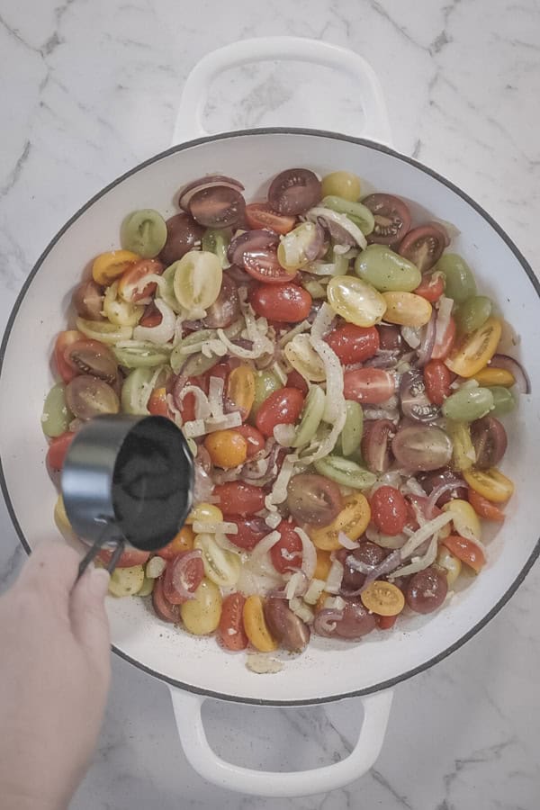 pouring water into a baking dish with cherry tomatoes, fennel, garlic and onions