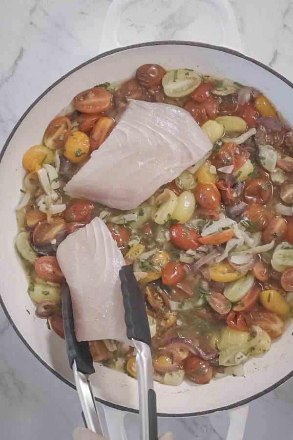 a pair of tongs is seen placing a raw fish fillet into a white round baking dish, filled with roasted tomatoes and fennel.