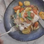 a blue plate with a piece of garlic toast, topped with flaky white fish, roasted cherry tomatoes, onions and fennel. A knife and fork are cutting the fish for eating.