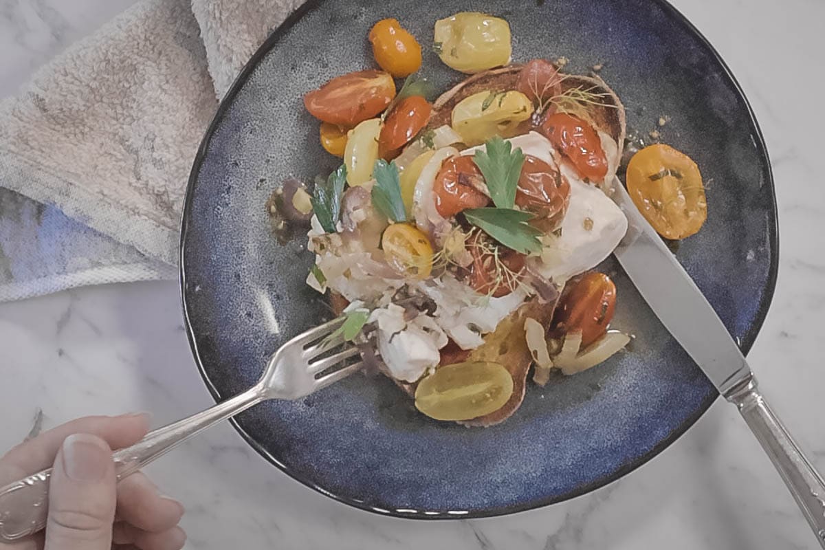 a blue plate with a piece of garlic toast, topped with flaky white fish, roasted cherry tomatoes, onions and fennel. A knife and fork are cutting the fish for eating.