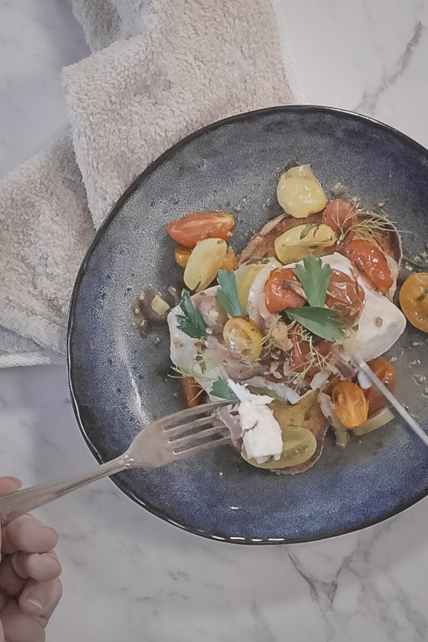 a blue plate with a beige napkin next to it. Silver cutlery is being held in a person's hand, forking up mouthfuls of flaky roasted fish with cherry tomatoes. 