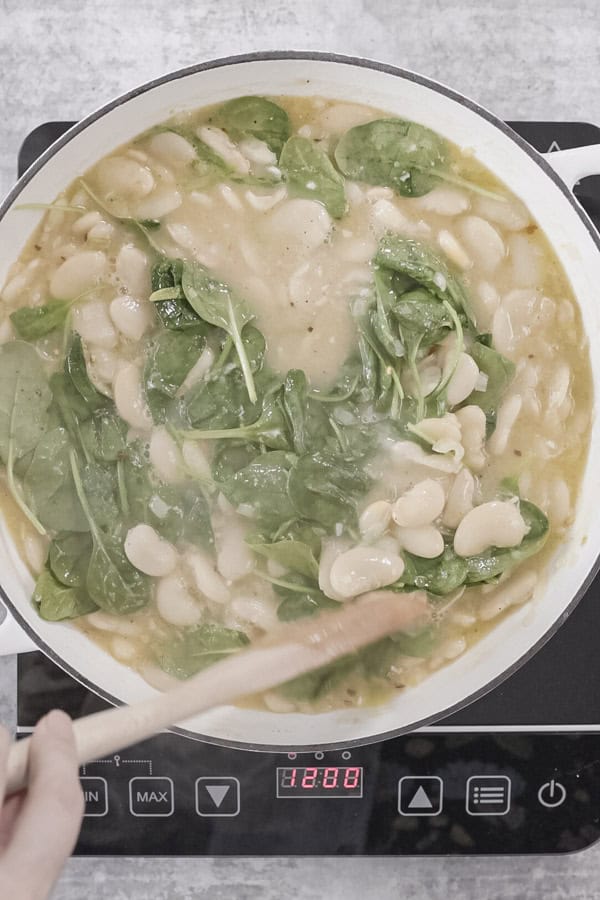 A hand stirs butter beans, sauce and spinach in a white pot.