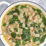 A large white cast iron pot is viewed from above. In the pot is creamy butter beans with lemon, garlic and spinach. Sprinkled on top is a scattering of red pepper flakes.