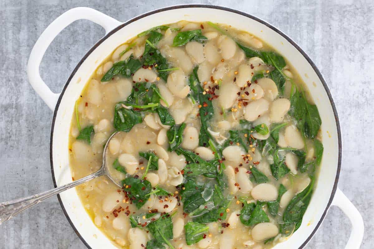 A large white cast iron pot is viewed from above. In the pot is creamy butter beans with lemon, garlic and spinach. Sprinkled on top is a scattering of red pepper flakes.
