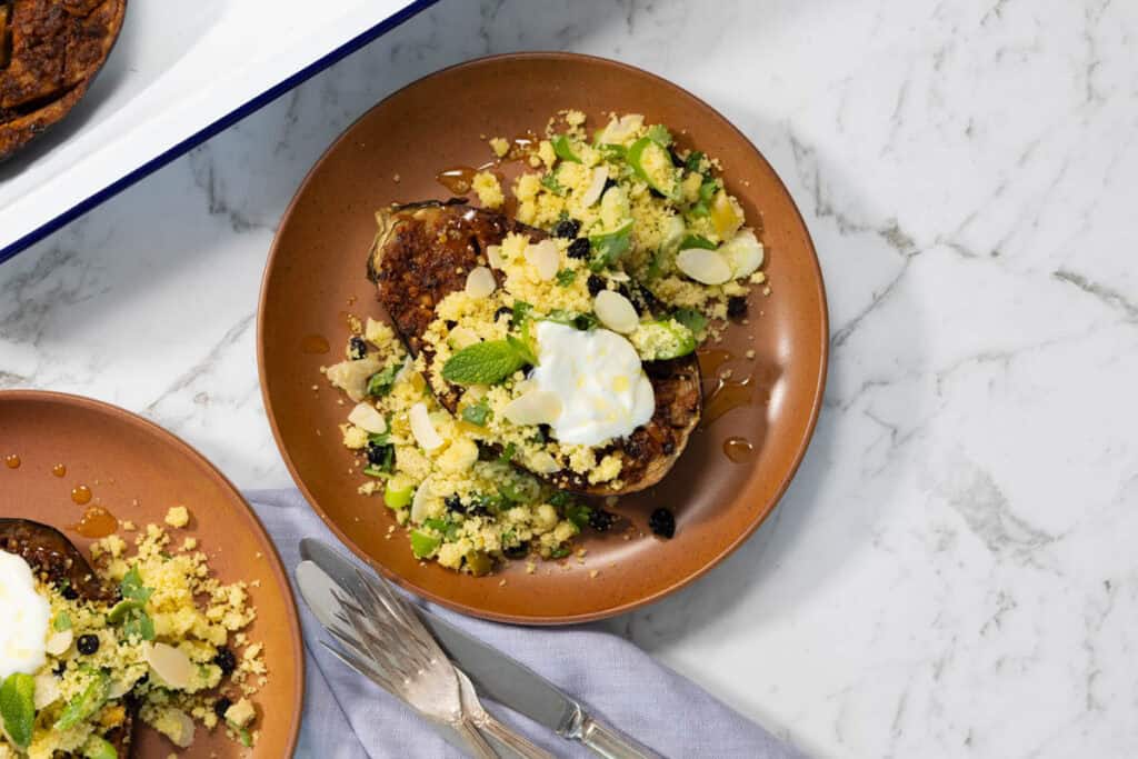 Landscape view of a terracotta plate holding a roasted eggplant half topped with yellow couscous, sliced green chilies, yogurt, and a drizzle of honey on a marble surface.