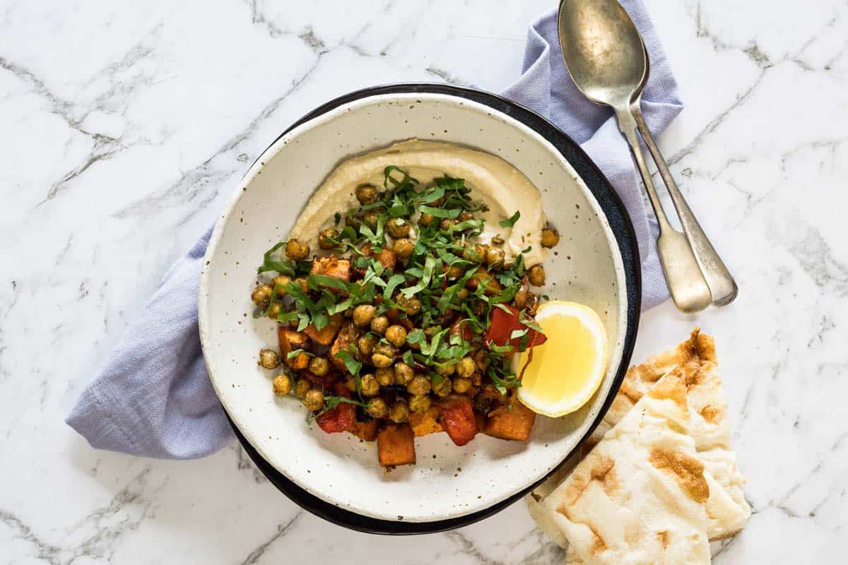 shallow ceramic bowl lined with hummus and topped with roasted sweet potatoes, peppers, crispy chickpeas, fresh chopped parsley, and a lemon wedge alongside pita bread.