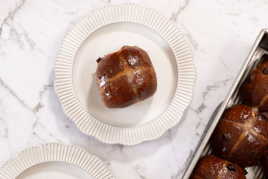 A single chocolate milk bread hot cross bun, on a white scalloped plate. The remaining tray of buns can be seen at the edge of frame.