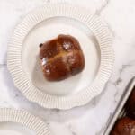 A single chocolate milk bread hot cross bun, on a white scalloped plate. The remaining tray of buns can be seen at the edge of frame.