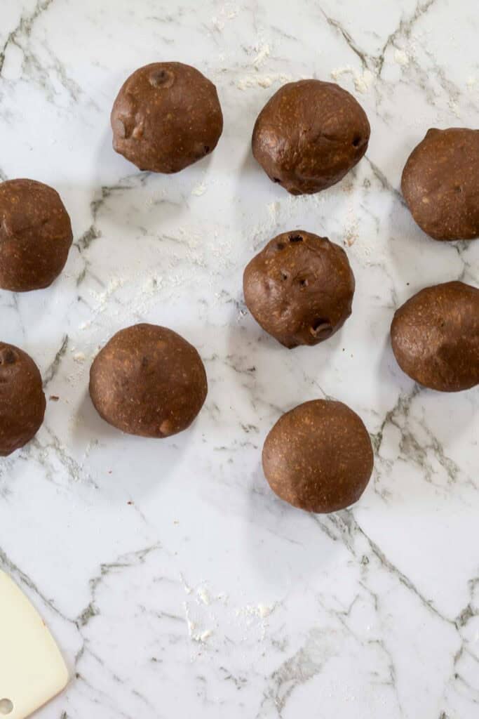 Balls of chocolate colored dough, being prepared to make chocolate milk bread hot cross buns. The dough balls are on a marble benchtop. 