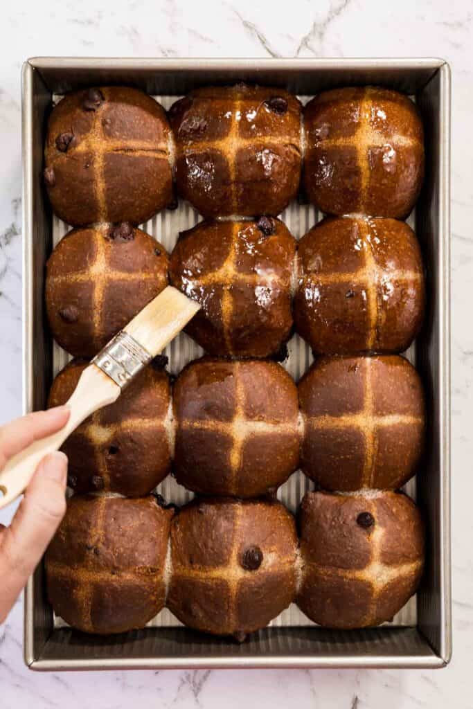 Brushing glaze over chocolate milk bread hot cross buns using a wooden pastry brush.