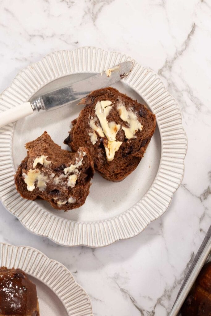 A split chocolate milk bread hot cross bun, sitting on a scalloped white plate, spread with butter and with a white handled knife on the side of the plate.