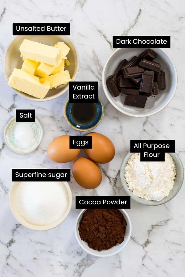 Overhead view of butter sticks, dark chocolate chunks, eggs, cocoa powder, white flour, sugar, and vanilla extract in bowls on a marble surface for chocolate and peanut butter brownies.
