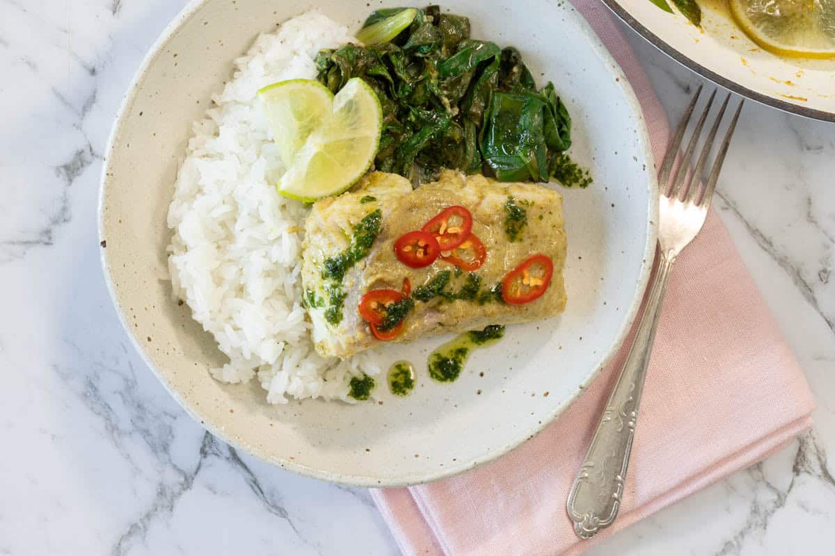 Overhead landscape shot of a ceramic bowl filled with coconut curry steamed fish, rice, Swiss chard, and lime with a fork on a pink napkin for coconut steamed fish.