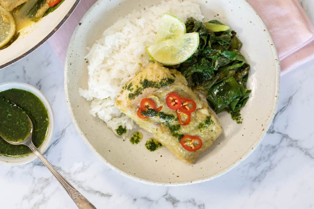 Overhead horizontal shot of a served bowl containing coconut steamed fish garnished with red chilis and herb oil, alongside white rice, steamed Swiss chard, and lime wedges for coconut steamed fish.