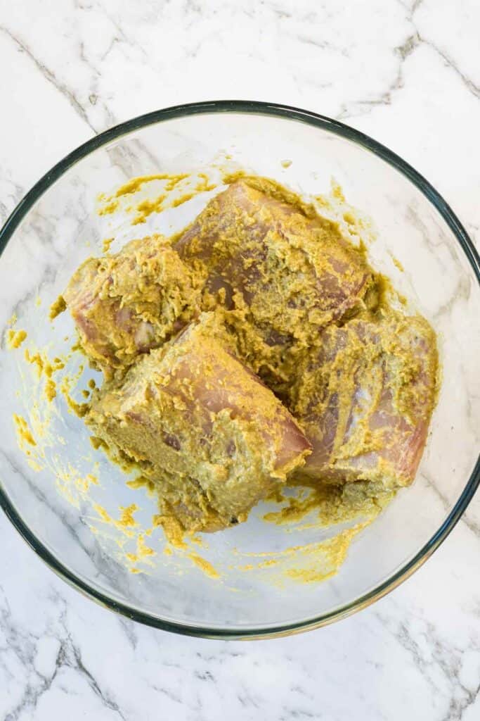 Top-down view of raw white fish fillets completely coated in a thick, green-yellow coconut curry sauce inside a glass mixing bowl for coconut steamed fish.