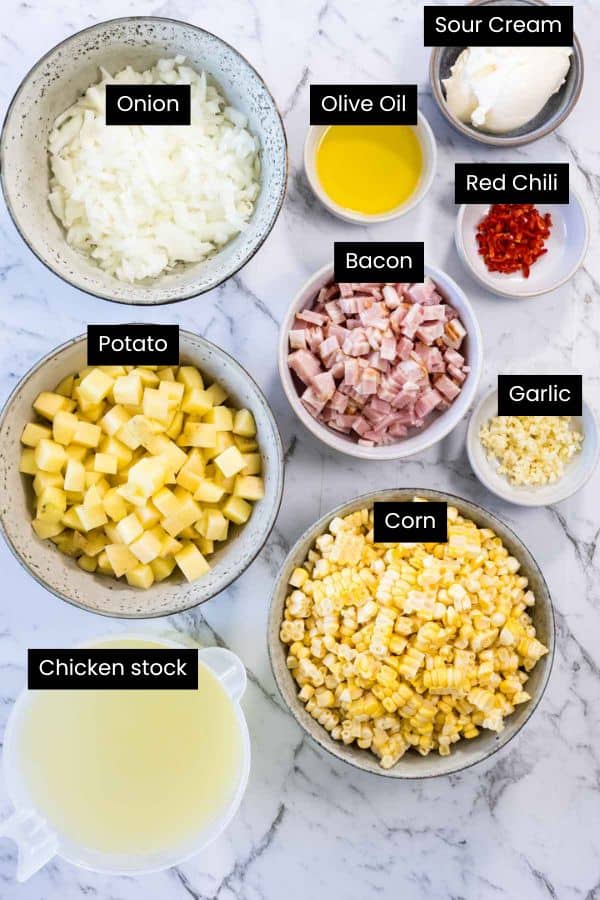 An overhead view of prepped ingredients in separate bowls on a marble surface, including fresh sweet corn kernels, diced potatoes, chopped bacon, minced onions, minced garlic, broth, olive oil, and cream cheese for a corn and bacon chowder.