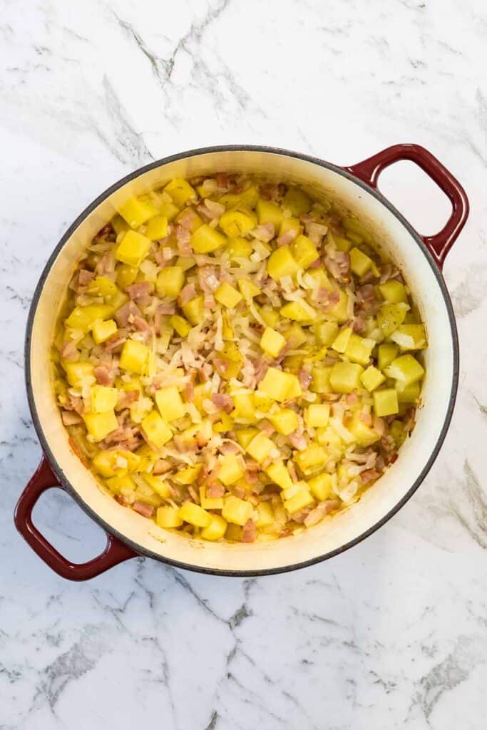 Cooked diced potatoes, translucent onions, and lightly rendered bacon pieces are stirred together inside a red enameled Dutch oven for corn and bacon chowder.