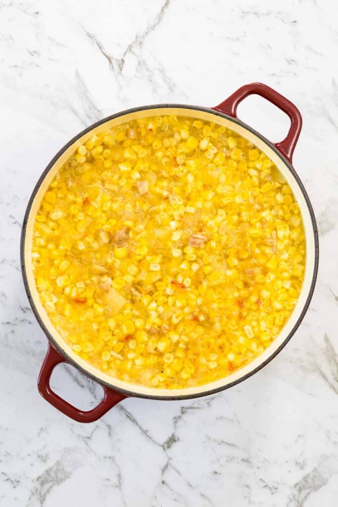 The corn and bacon chowder is mixed together thoroughly and simmers directly to the rim inside the red enameled Dutch oven on a white marble countertop for corn and bacon chowder.